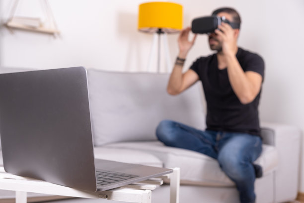 年轻人戴着VR眼镜，戴着耳机在家里用笔记本电脑，未来科技，教育视频游戏图片下载