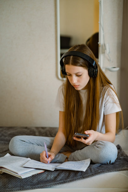 一个十几岁的女孩坐在家里的床上，戴着耳机和智能手机，准备上课。图片下载