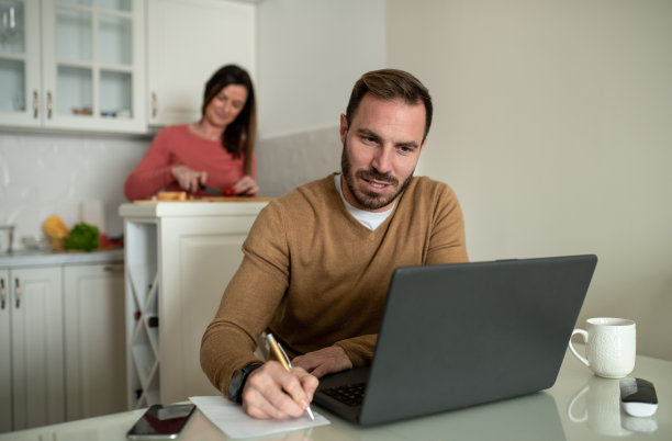 一个年轻的自由职业者正坐在餐厅里工作，而他的妻子在他身后的厨房里准备饭菜。图片下载