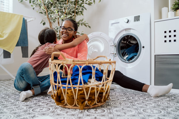 快乐的女人和女儿一起做家务，他们一起整理衣服，把五颜六色的东西扔进洗衣机，小女孩拥抱妈妈，感谢她的帮助图片下载