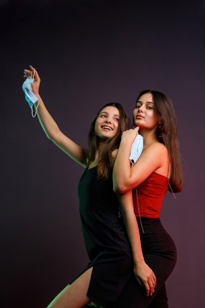 侧面的照片，两个性感的女孩穿着晚礼服，快乐和严肃的表情，拿着医用口罩摆姿势对着镜头在流行病时间。图片下载