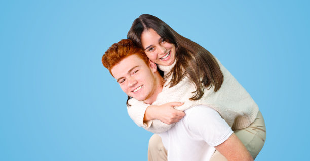 两个相爱的年轻人玩得很开心，相当快乐可爱的微笑女孩是坐在她男朋友的背部孤立的蓝色背景。图片下载