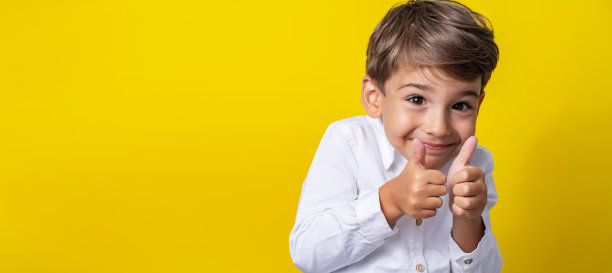 前视图是一个四岁的白人小男孩，站在黄色背景的工作室前微笑着，举着大拇指支持孩子和成功的概念图片下载