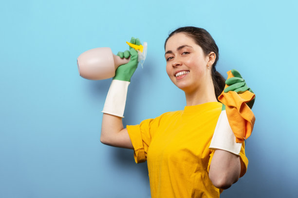 一个面带微笑的白人年轻女子，戴着橡胶手套，手里拿着喷雾器和抹布。蓝色背景与复制空间。清洁服务的概念图片下载