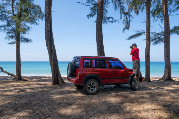 年轻的亚洲男性旅行者和摄影师站在四轮驱动的汽车上，拍摄泰国普吉岛美丽的海滩旅游摄影概念图片下载