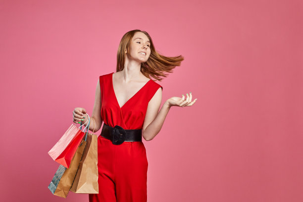 年轻女子带着幸福积极的微笑，穿着时髦的红色衣服，兴奋地拿着购物袋在粉红色的背景上出售。图片下载