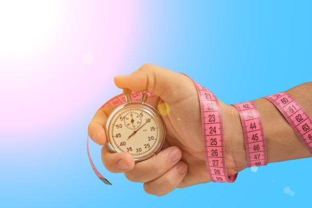用卷尺包着的手握秒表，蓝色背景，明亮的阳光。健身运动和健康。图片下载