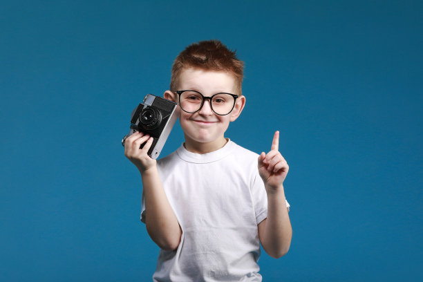 小男孩正在用老式相机拍照，手指向上。蓝色背景上的小男孩拿着老式相机。旧技术的概念。孩子学习摄影图片下载