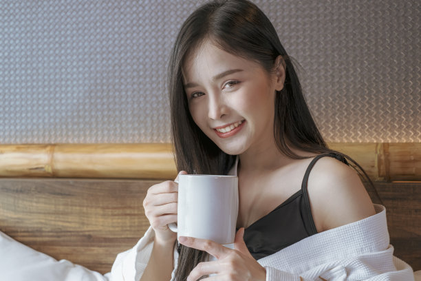 快乐美丽的年轻亚洲妇女放松地喝和看着一杯热咖啡或茶在早上的床上。图片下载