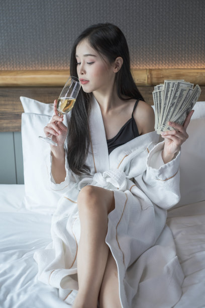 美丽的女人穿着浴袍，在卧室里喝着香槟。年轻女子躺在床上喝香槟。图片下载