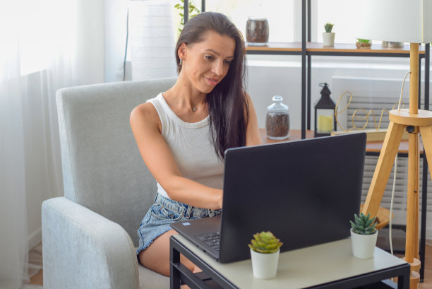 一个年轻快乐的女人坐在书桌前用电脑工作的大头照。愉快迷人的微笑女士看着笔记本电脑屏幕，购物，社交网络，在线学习远程。图片下载