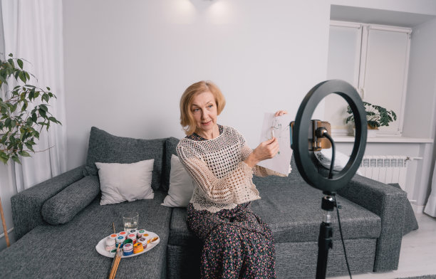 这位艺术家是一位上了年纪的妇女，她正在为一个博客展示用摄像机绘制的画笔。绘画，在家画画，三脚架上的相机，一个绘画和诉说的博主。图片下载