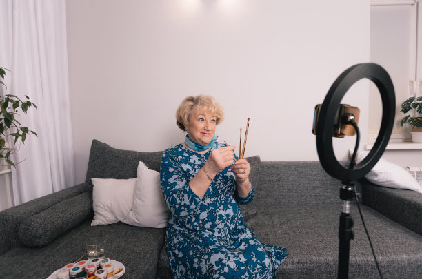 这位艺术家是一位上了年纪的妇女，她正在为一个博客展示用摄像机绘制的画笔。绘画，在家画画，三脚架上的相机，一个绘画和诉说的博主。图片下载