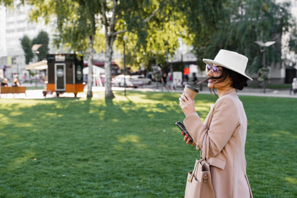 拿着智能手机和咖啡杯的博主的街头风格照片。女孩戴着白色的帽子，穿着白色的连衣裙和夹克。图片下载