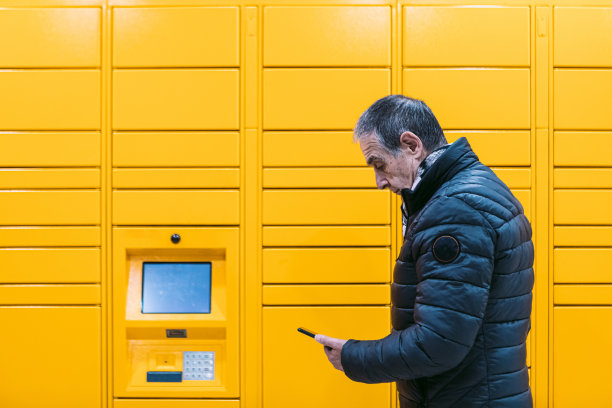 穿灰色外套的男人看着他的手机，从黄色储物柜里拿起一个包裹。消息传递概念，比较在线，电子商务和包裹图片下载