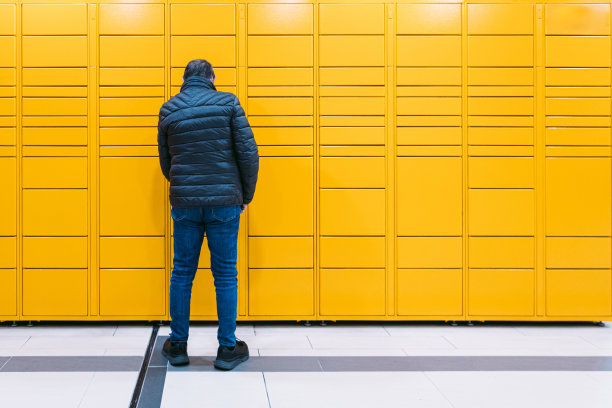 后面的男子扫描手机上的代码，从黄色储物柜里取走一个包裹。消息传递概念，比较在线，电子商务和包裹图片下载