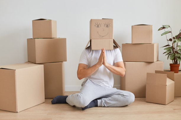 在室内拍摄的这张照片中，一名不知名的男子穿着白色t恤，坐在纸板箱旁边的地板上，把纸板箱顶在头上摆姿势，在搬新家期间，他试图通过瑜伽练习放松自己。图片下载