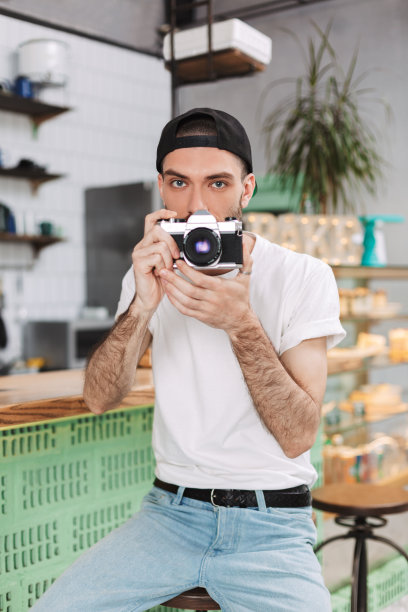 穿着白t恤，戴着黑帽子的年轻人坐在吧台前，用小相机遮住自己的脸，在咖啡馆里消磨时间图片下载