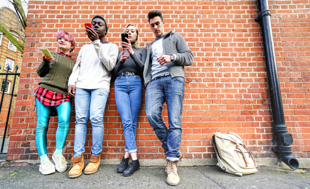 在伦敦肖尔迪奇区，多文化的城市朋友们在墙壁上使用移动智能手机-技术上瘾的生活方式概念，无聊的男孩和女孩在智能手机上发短信-明亮温暖的过滤器图片下载