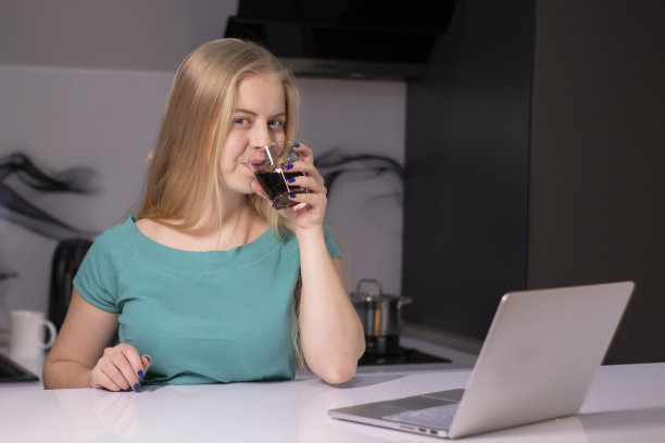 漂亮的金发女孩，穿着绿色的t恤，在灰色的房间里，在笔记本电脑旁工作或学习，手里拿着一个装有棕色液体饮料的玻璃杯。咖啡，茶，酒精，威士忌，果汁图片下载