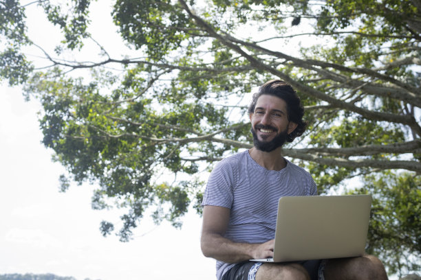 一个阳光明媚的日子里，一个年轻人在公园里，腿上放着笔记本电脑。图片下载