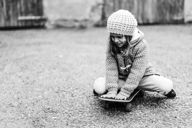 穿着冬装的可爱小女孩坐在户外后院的滑板上-微笑的小女孩玩滑板-复古的黑白版图片下载