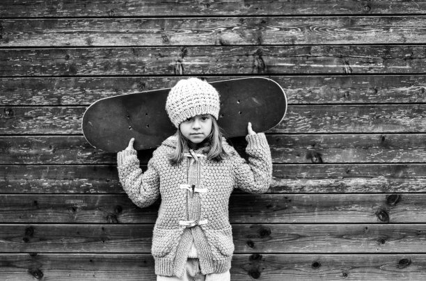 白人小女孩滑板运动员的肖像，穿着羊毛冬装，肩上扛着滑板，看着木制背景下的相机-童年和运动概念-黑白图片下载