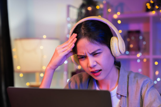 年轻的亚洲女性，戴着耳机，坐在椅子上，桌上放着一堆文件，晚上看着电脑屏幕。图片下载