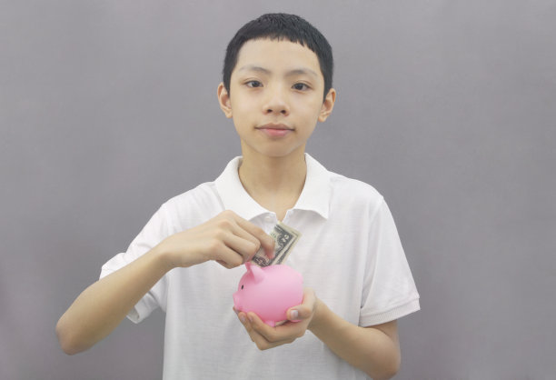 正面，英俊和友好的亚洲男孩的肖像，在灰色的背景上持有美元和储蓄罐。图片下载