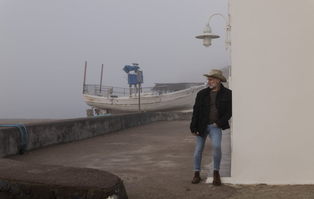 戴着牛仔帽和牛仔裤的成年男子的肖像靠墙，背景是渔船。西班牙阿尔梅里亚图片下载