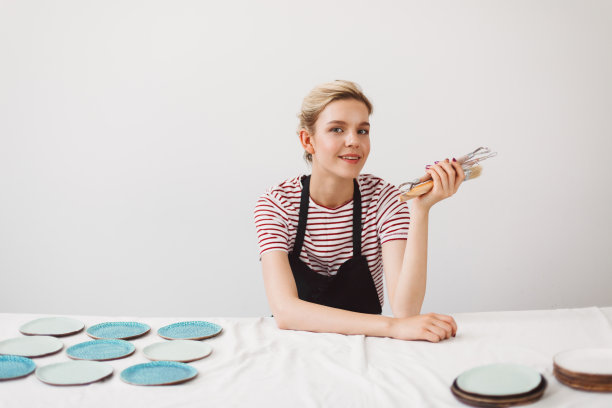 穿着黑色围裙和条纹t恤的漂亮女孩坐在桌子旁，手里拿着盘子和陶艺工具，梦幻般的看着陶艺工作室的镜头图片下载