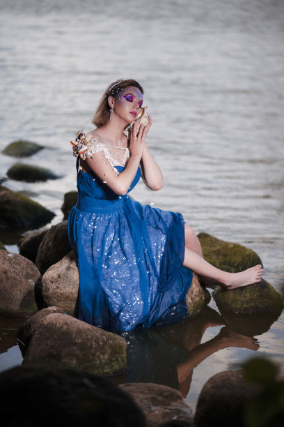 放松积极的女士在艺术的湿蓝色连衣裙和脸上装饰着Strasses拿着海贝坐在户外的岩石海岸。图片下载