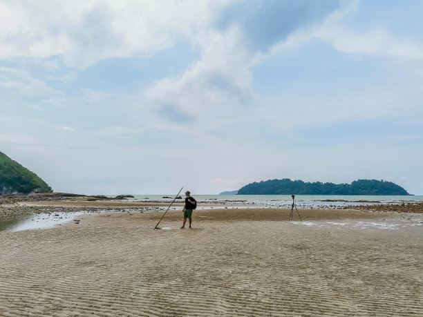 男性背包客站在低潮的沙滩上与岛屿和多云的天空背景。寻找灵感的摄影师。沙滩上的三脚架。旅行生活冒险概念图片下载