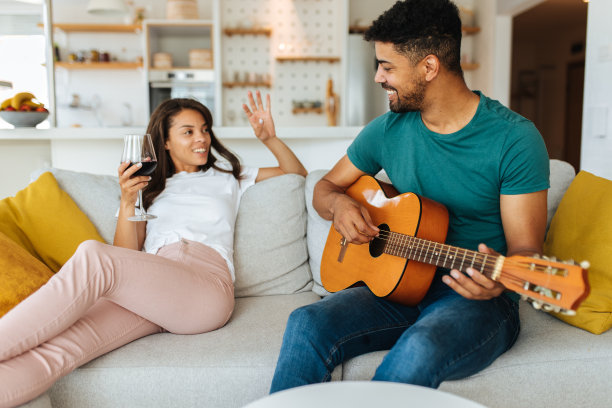 这对非裔美国夫妇在一起度过了浪漫的时光。这个男人正在和他心爱的女朋友弹吉他。一个女人坐在他旁边的沙发上喝着红酒。图片下载