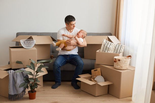 这是一名男子的照片，他穿着白色休闲t恤和牛仔裤，和他的宝贝女儿坐在沙发上，在搬家后打开箱子，试图让哭泣的孩子安静下来。图片下载