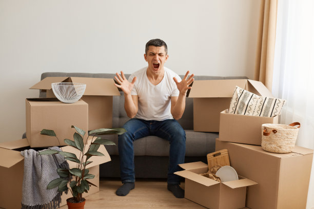 照片中，一名愤怒的男子穿着白色t恤和牛仔裤，坐在沙发上，周围都是纸箱，他举起手臂，咄咄逼人地尖叫着，移动和转移。图片下载