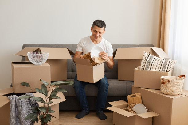 照片中的男子穿着白色t恤和牛仔裤，微笑着坐在沙发上，周围是纸箱，拿着盘子，在搬家后打开包装。图片下载