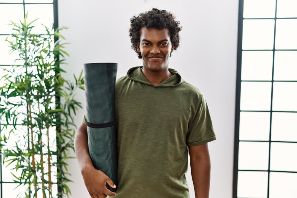 一头卷发的非洲男子拿着瑜伽垫在工作室里，脸上带着幸福和凉爽的微笑。幸运的人。图片下载