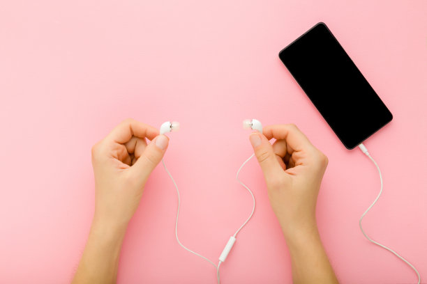 年轻的成年女性手握白色的小耳机，在浅粉色的桌子背景上听智能手机上的音乐。柔和的颜色。特写镜头。黑屏上空空如也。自顶向下的观点。图片下载