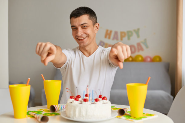 照片中，一个乐观的年轻人穿着白色休闲t恤，面带微笑，手拿蛋糕坐在餐桌旁，手指对着镜头，邀请你参加他的生日派对。图片下载