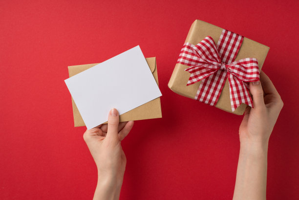 第一人称俯视图照片的情人节装饰女孩的手握信件信封和工艺纸礼盒与方格丝带蝴蝶结在孤立的红色背景与空白空间图片下载