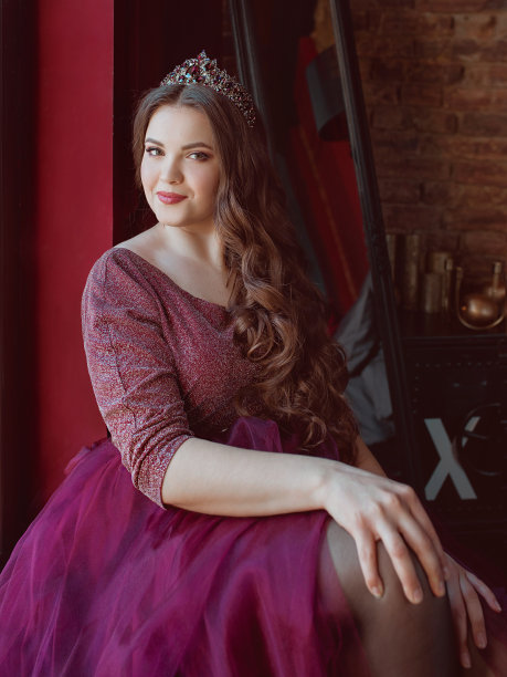 美丽的年轻漂亮可爱的公主公主穿着紫色的长裙和皇冠，留着长发，化妆室内在极简的阁楼室内坐在窗边图片下载