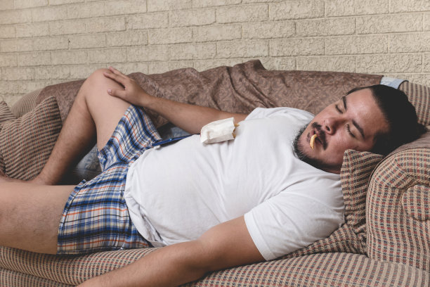 一个亚洲和美国血统的又懒又累的人在沙发上打盹，嘴里还叼着一根薯条。图片下载