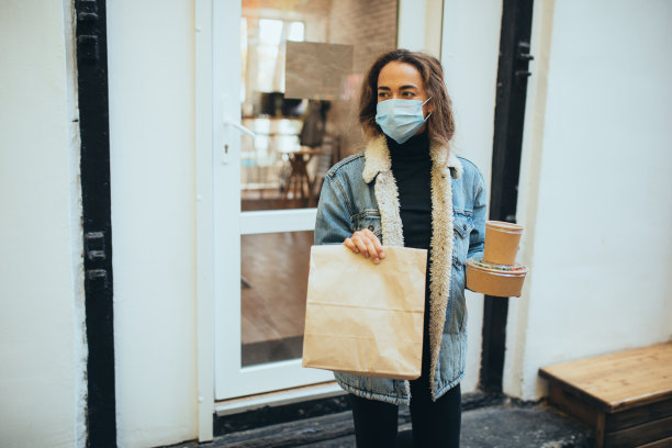 年轻女子戴着防护口罩，拿着装着食物和碗的外卖纸袋，站在餐厅窗户旁。隔离期间的社会距离导致了大流行。图片下载