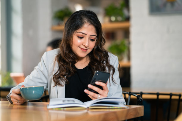 一名年轻女子一边使用智能手机，一边在咖啡馆看书、喝咖啡。图片下载