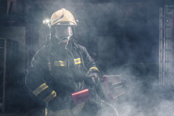 一名勇敢的消防队员自信地站着，穿着全套防护装备，出场和头盔。黑色背景与烟和蓝光。图片下载