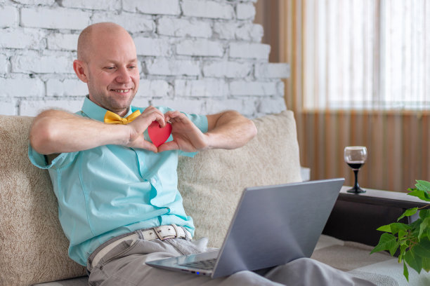 爱在网上。距离是爱的宣言。一个快乐的男人在情人节独自在家。一位优雅的成年男子在浪漫的约会中手持红心形状的卡片图片下载
