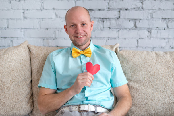 爱在网上。距离是爱的宣言。一个快乐的男人在情人节独自在家。一位优雅的成年男子在浪漫的约会中手持红心形状的卡片图片下载