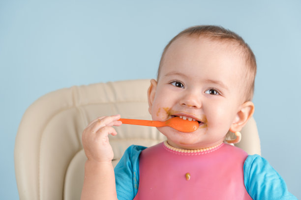 12-17个月大、脏兮兮的新生婴儿戴着粉色围嘴，面带微笑，把一个勺子放进嘴里，用牙齿咬它图片下载