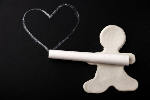 一个小小的白色橡皮泥人，手里拿着粉笔，在一块黑色的板子上画着一个白色的心的轮廓图片下载